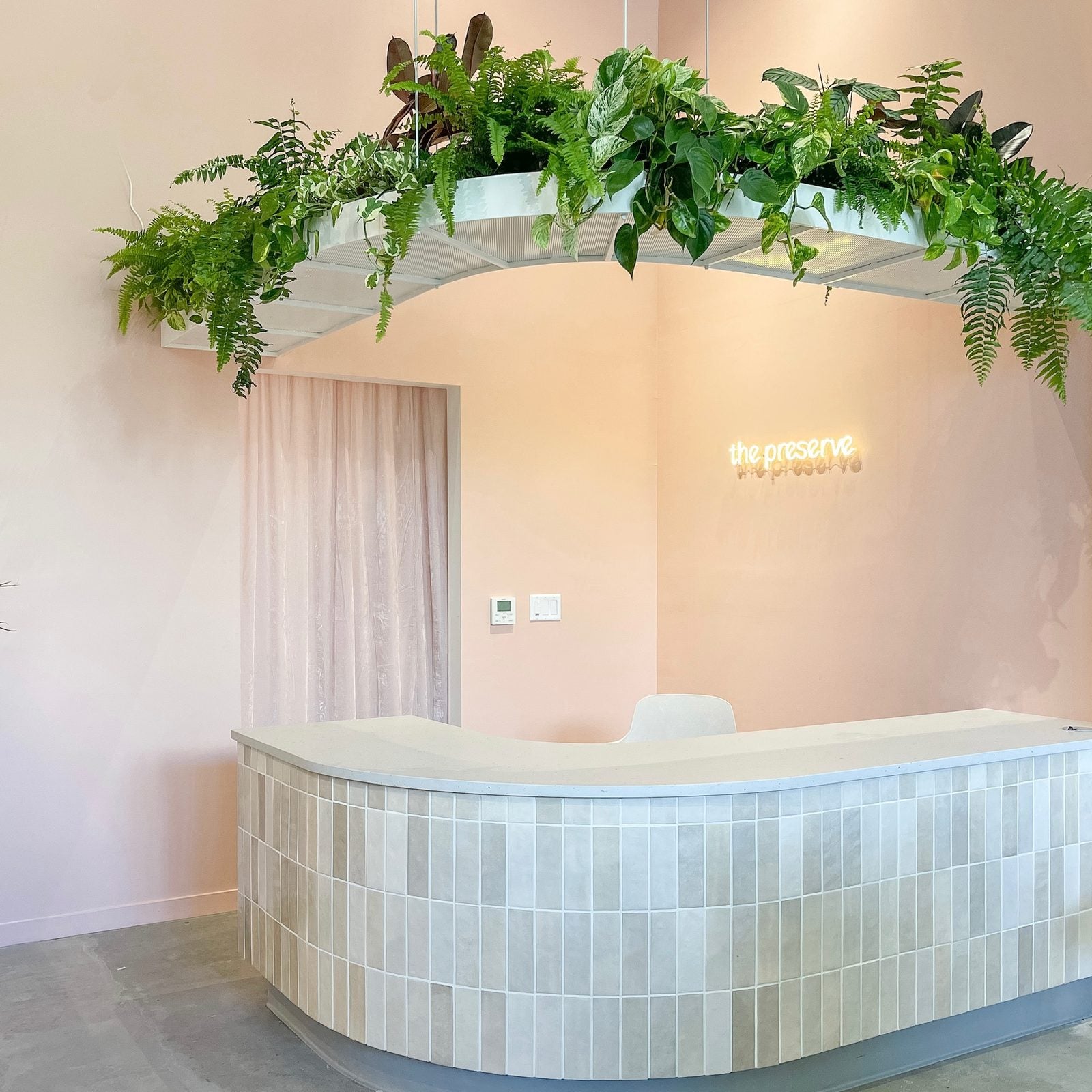 The Preserve's reception desk beneath an arch of ferns and pothos, with a neon sign on a peach wall
