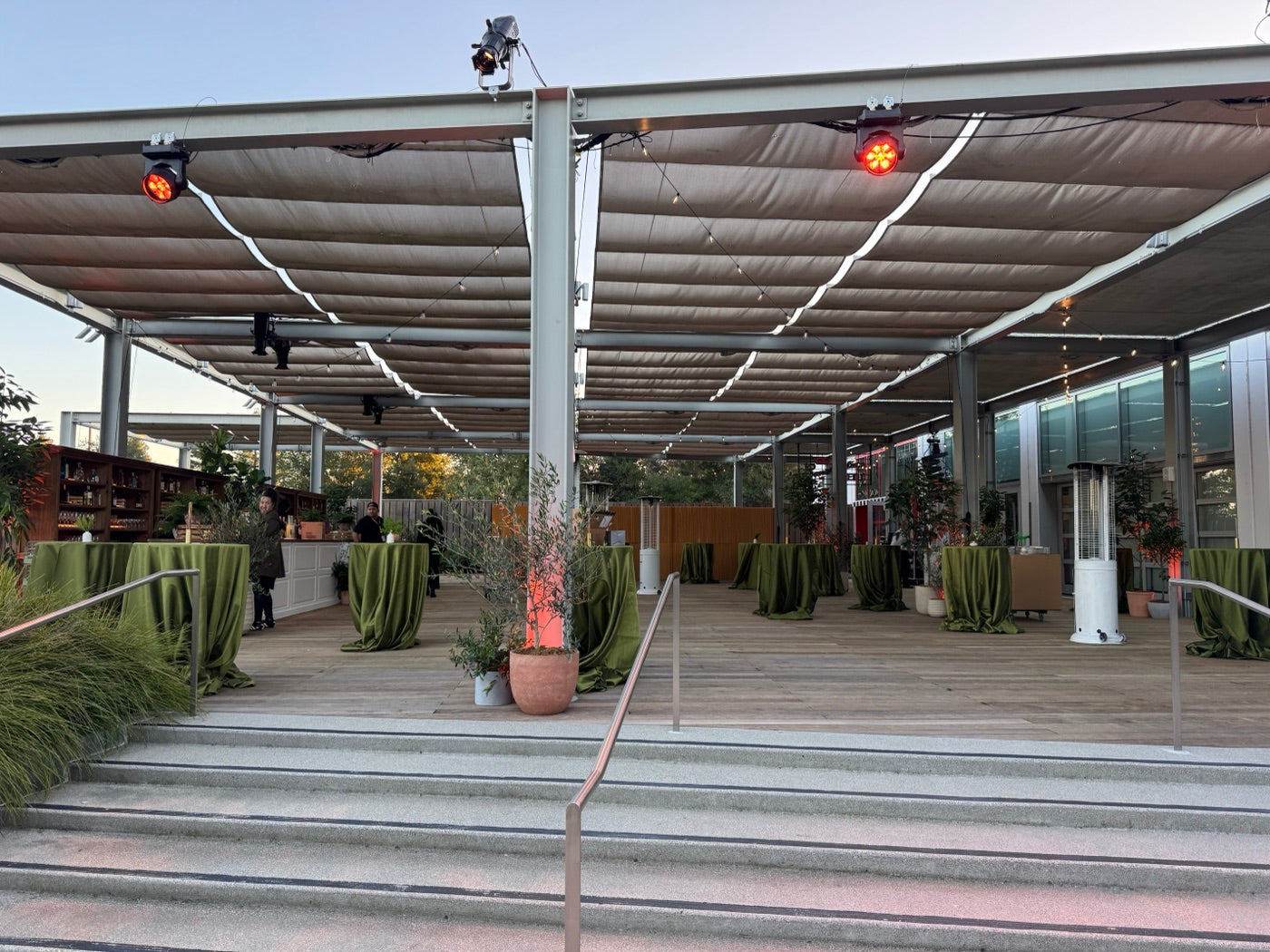 Potted trees surround green-draped cocktail tables and an outdoor bar on a covered deck.